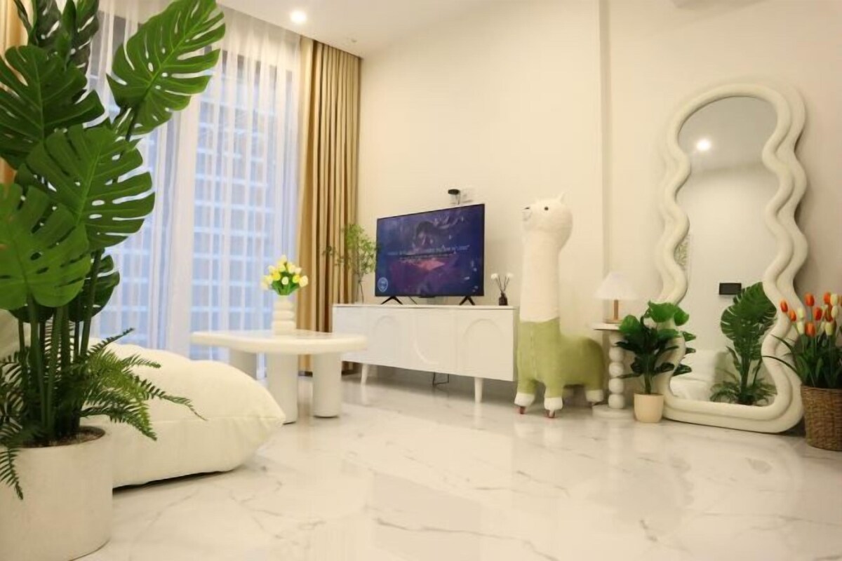 Ein modernes Wohnzimmer mit einem weißen Sofa, einem Fernseher auf einem Schrank, einem weißen Schränkchen und verschiedenen Pflanzen.