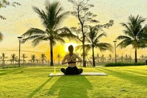 A person sitting on a mat in a grassy area with palm trees and a sunset in the background. - Hera House Vinhomes Grand Park Ho Chi Minh City (Ho Chi Minh City)