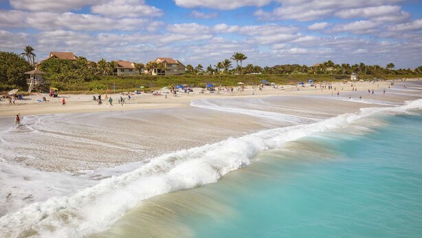A beach with a wide sandy shore, clear turquoise water, and a few buildings in the background.