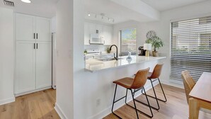 A modern kitchen with white cabinets, a built-in microwave, and a bar area with two stools.