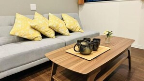 Ein Wohnzimmer mit grauem Sofa, gelb gemusterten Kissen, einem Holztisch mit Teekannen und einer kleinen Vase mit Blumen.