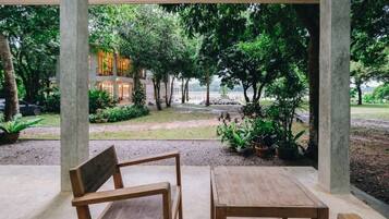 A covered outdoor seating area with wooden furniture, surrounded by lush greenery and a view of a building in the background.