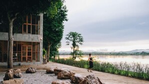 A serene outdoor area with large rocks, a modern building with large windows, and a view of a lake and distant hills. - Loogmai Guesthouse (Chiang Khan)