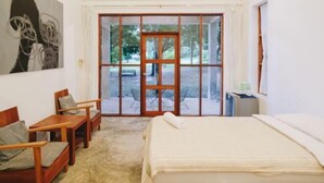 A hotel room with a bed, two wooden chairs, a small table, a painting on the wall, and a sliding glass door leading to an outdoor area. - Loogmai Guesthouse (Chiang Khan)