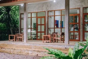 Un espace extérieur abrité, doté de chaises et de tables en bois, de grandes portes vitrées et d’une structure en béton.
