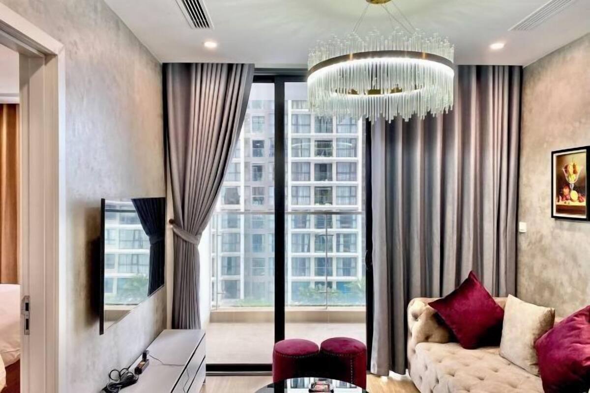 A modern living room with a large window, a chandelier, a sofa with cushions, and a flat-screen TV mounted on the wall.
