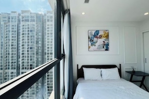 A hotel room with a large window, a bed with white bedding, a small table, and a colorful abstract painting on the wall.
