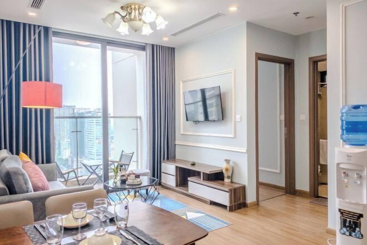 A modern living room with a sofa, coffee table, and dining table set up. There is a TV mounted on the wall, a water dispenser, and a large window with curtains.