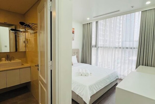 A hotel room with a bed, a bedside table, a window with curtains, a bathroom with a sink and mirror, and a towel rack.