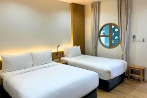 Habitación de hotel con dos camas, una mesita de noche pequeña, un ventanal redondo con vistas y una lámpara fijada a la pared.