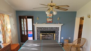 Une pièce avec une table de ping-pong, une cheminée, un ventilateur de plafond et un fauteuil en osier.