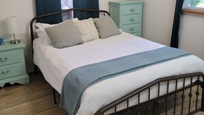 Un lit bien fait, recouvert d’une couvre-lit blanche et bleue, avec une tête de lit en bois et une table de chevet surmontée d’une lampe.