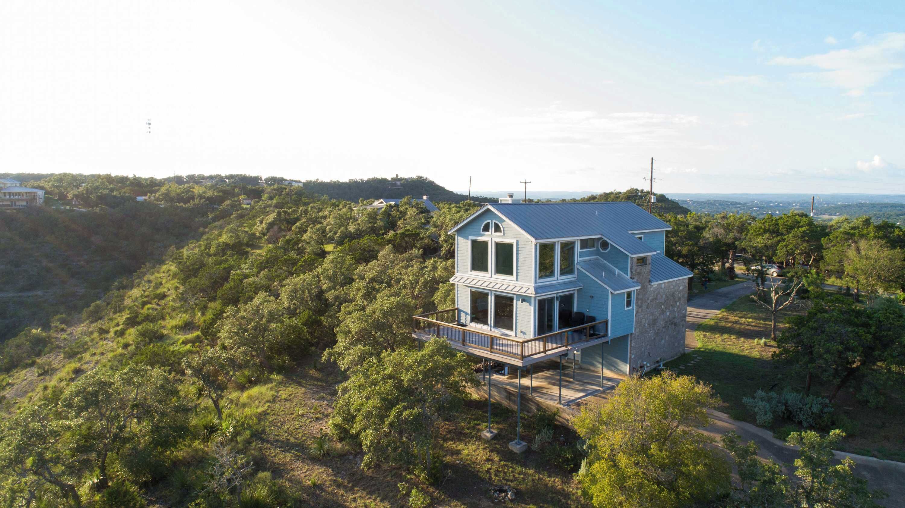 A two-story house with a balcony, surrounded by lush greenery and a hilly landscape.
