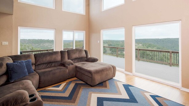 A spacious living room with a large sectional sofa, a ceiling fan, and large windows offering a view of a balcony with a wooden railing. - Cliffside at Canyon Lake - Monthly Rental (New Braunfels)