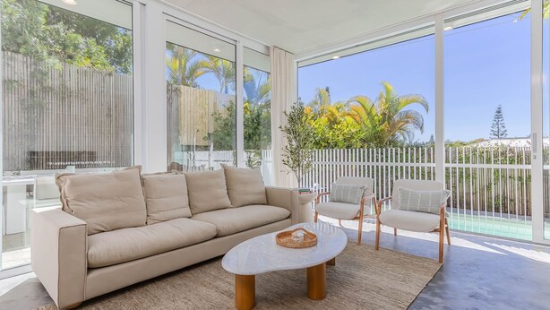 A modern living room with a beige sofa, two armchairs, and a coffee table. Large windows offer views of palm trees and a white fence. - Salt & Stone Luxe Hideaway Byron Bay (Byron Bay)