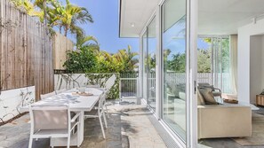 Une terrasse extérieure moderne avec une table et des chaises blanches, une porte vitrée donnant sur un balcon et une clôture en bois agrémentée de verdure.