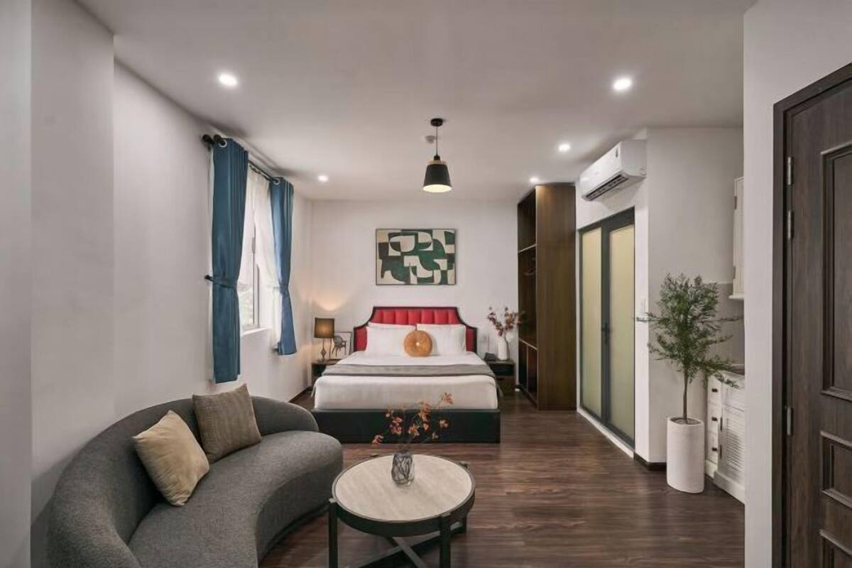 A modern hotel room with a curved sofa, a round coffee table, a large bed with a red headboard, and a wall-mounted abstract art piece.