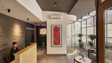A modern hotel lobby with a reception desk, a large floral artwork, a glass enclosure, and potted plants.