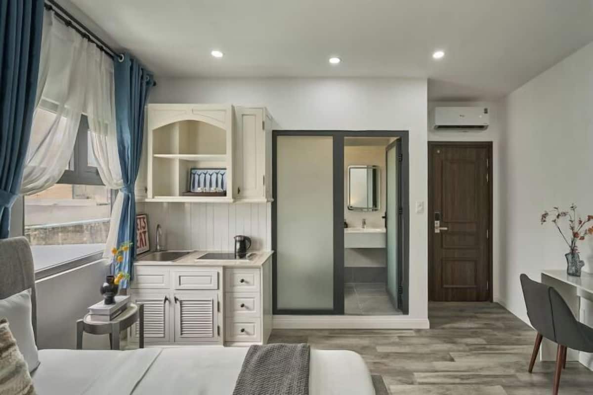 A bedroom with a bed, a bedside table, a window with curtains, a built-in wardrobe, a bathroom with a mirror and sink, and a door leading to another room.