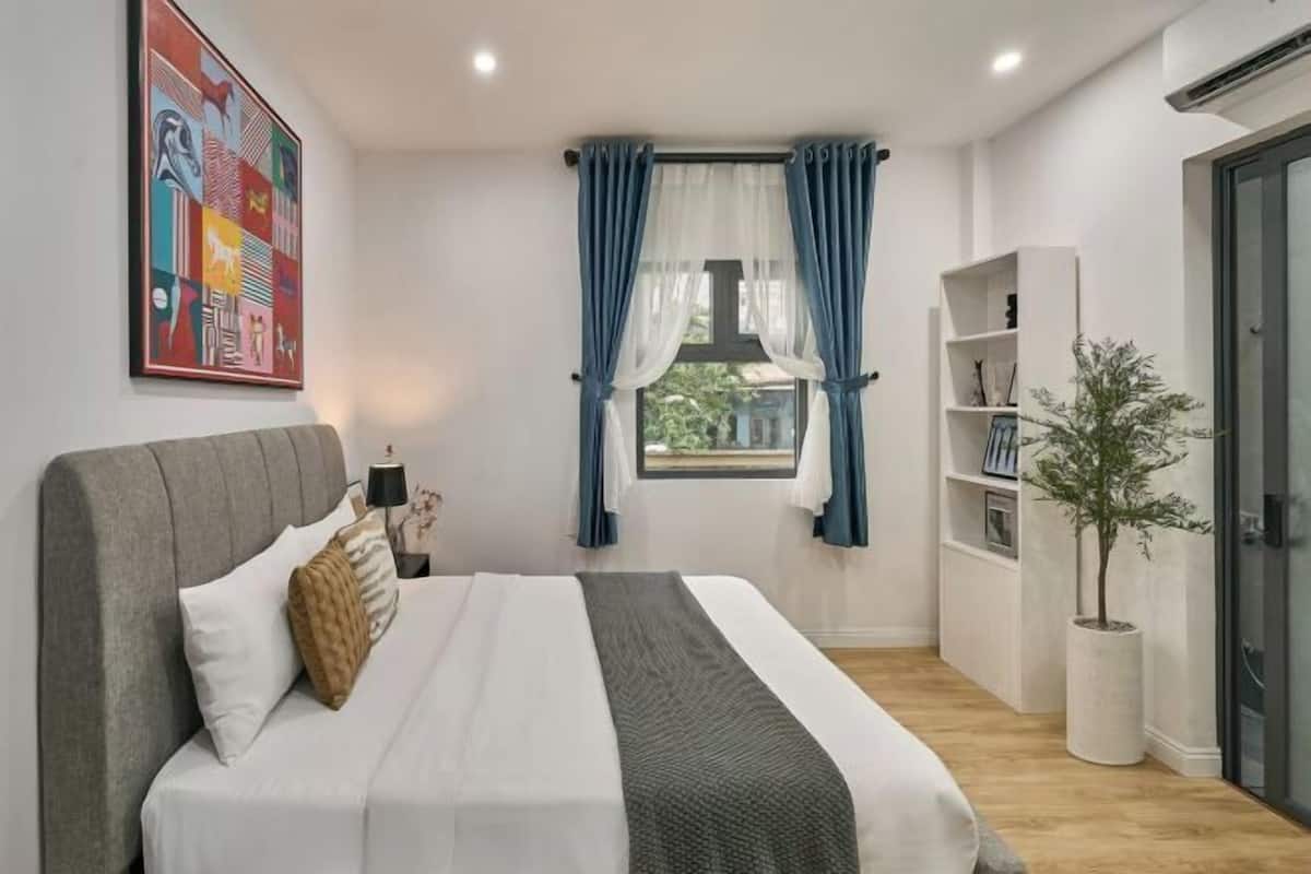 A bedroom with a bed, a window with curtains, a bedside table, a lamp, a bookshelf, and a potted plant.