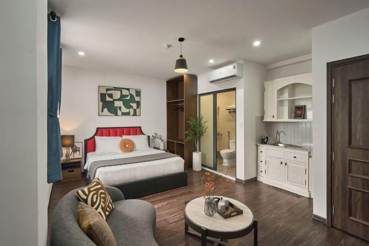 A modern hotel room with a bed, a seating area, a small table, a wardrobe, and a bathroom visible through a glass door.