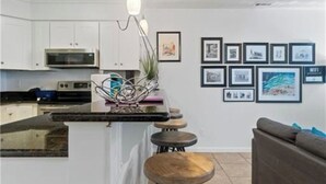 A modern kitchen with white cabinets, a microwave, and a bar area with stools. There are framed pictures on the wall and a gray sofa in the living area. - Vibe District, Atlantic Surf Park, VB Sports Center, Boardwalk & Oceanfront (Virginia Beach)