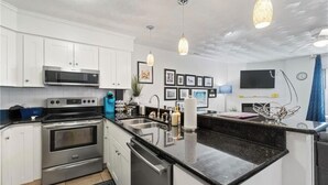 A modern kitchen with white cabinets, stainless steel appliances, a black granite countertop, pendant lighting, and a flat-screen TV mounted on the wall. - Vibe District, Atlantic Surf Park, VB Sports Center, Boardwalk & Oceanfront (Virginia Beach)