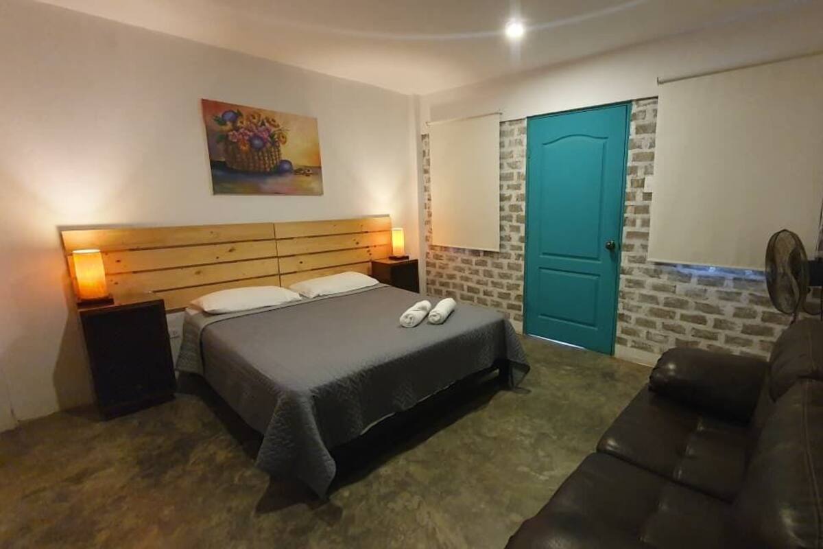 A bedroom with a bed, bedside lamps, a wooden headboard, a blue door, a fan, and a brown leather couch.