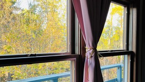 A window with a curtain tied in the middle, overlooking a blue deck and autumn foliage.