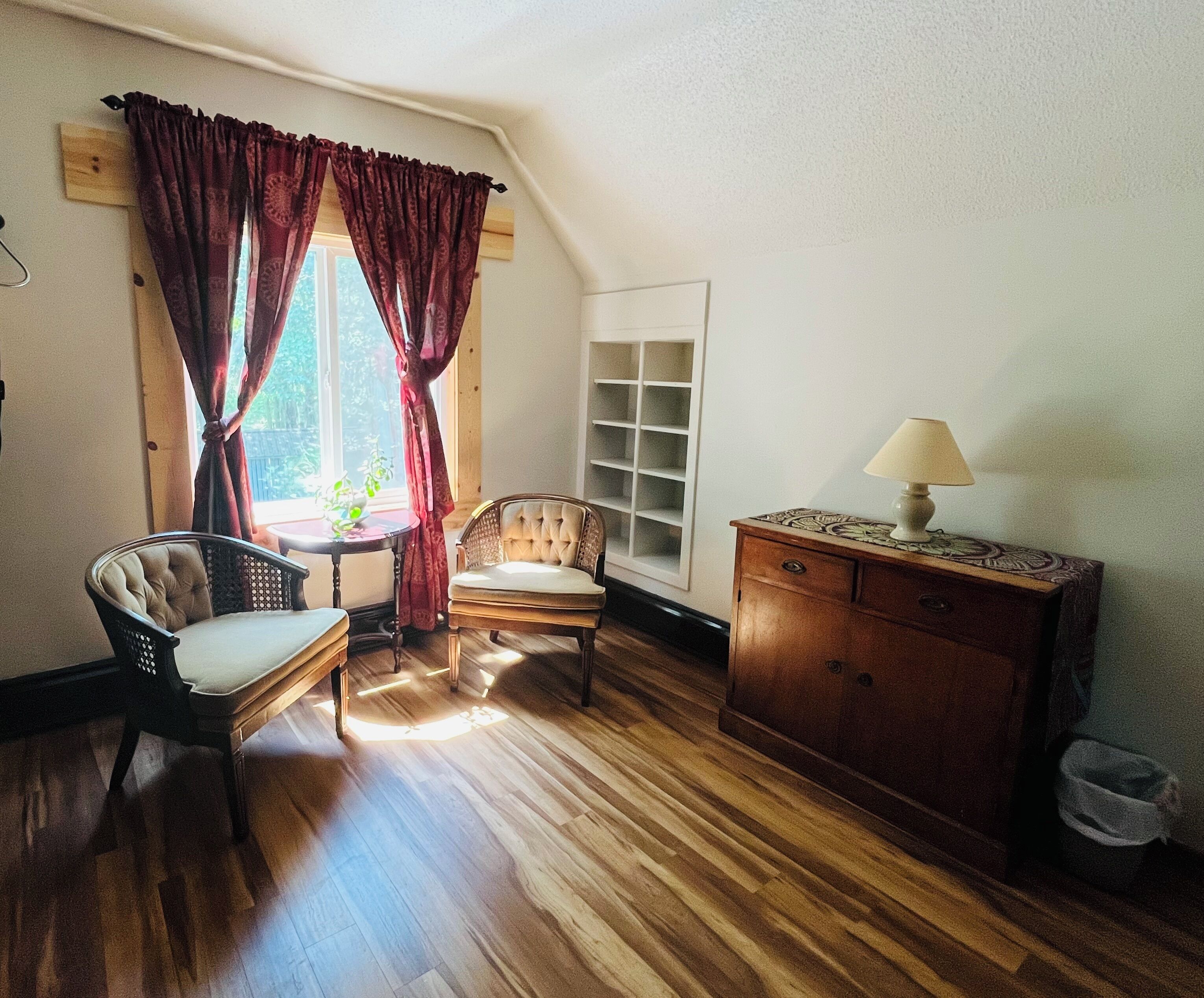 A room with a wooden floor, two chairs, a table, a lamp, a cabinet, and a window with curtains.