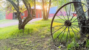 A rustic scene with a large wooden wheel, trees, and a red barn in the background.