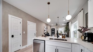 A modern kitchen with white cabinetry, a built-in dishwasher, a large island with a sink, pendant lighting, and a view of the living area. - The Boro Suite I 3BR Near Downtown & MTSU w/ Ping Pong (Murfreesboro)