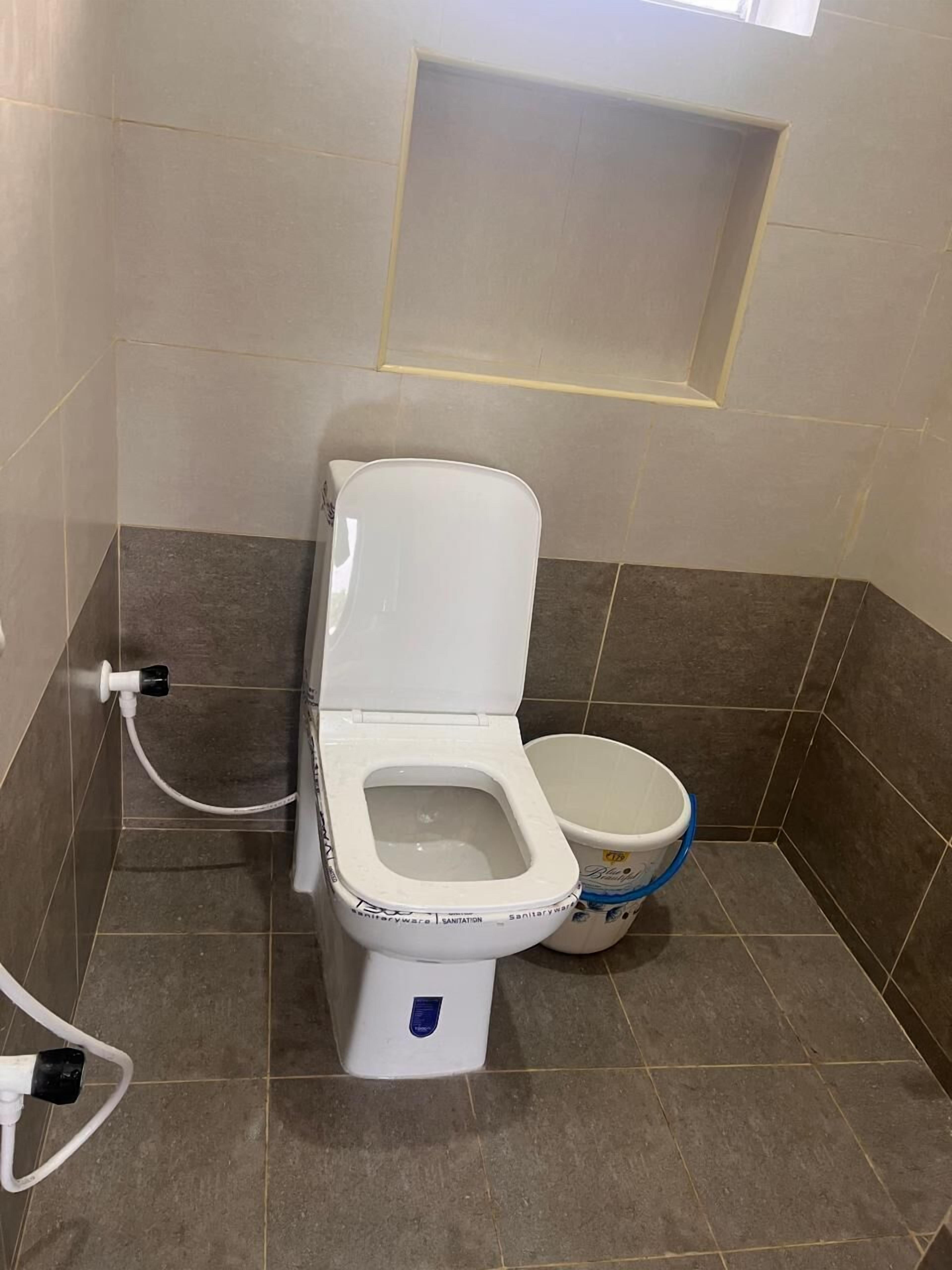 A toilet with a bidet attachment, a white bucket, and a small window above the toilet.
