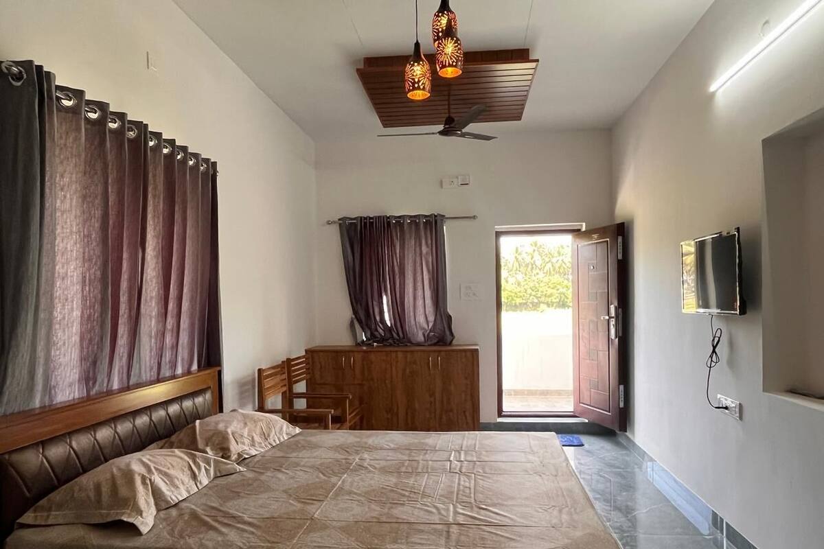 A bedroom with a bed, wooden headboard, curtains, a wooden bench, a ceiling fan, and a television on the wall.
