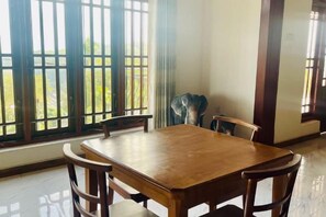 A dining area with a wooden table and chairs, large windows with a view of greenery, and a statue on the wall. - Villa Avora Ridge (Kandy)