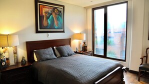 Une chambre avec un grand lit, deux tables de chevet avec des lampes, une œuvre d’art encadrée au-dessus du lit, et une vue à travers une porte-fenêtre coulissante.