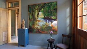 A room with a large painting of a forest scene, a statue on a cabinet, and a vase with flowers on a small table.