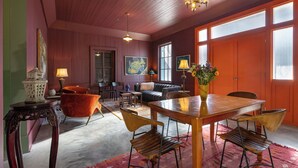 A spacious room with a wooden ceiling, a dining table with chairs, a sofa, and a large window with blinds.