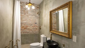 A bathroom with a brick wall, a white toilet, a black sink, a gold-framed mirror, and a shower area.