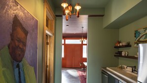 A narrow hallway leading to a kitchen with a dining area, a large painting on the left, and a wooden ceiling.