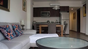 A modern living room with a grey sofa, colorful pillows, a glass-top coffee table, and a dining area with a wooden table and chairs. - Cozy Family Condo - Pool & terrace - 10 minute to the beach - Nuevo Vallarta (Nuevo Vallarta)