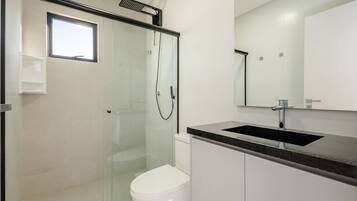 Un baño moderno con cabina de ducha de vidrio, inodoro blanco, lavamanos con encimera negra y un espejo grande sobre el lavamanos.