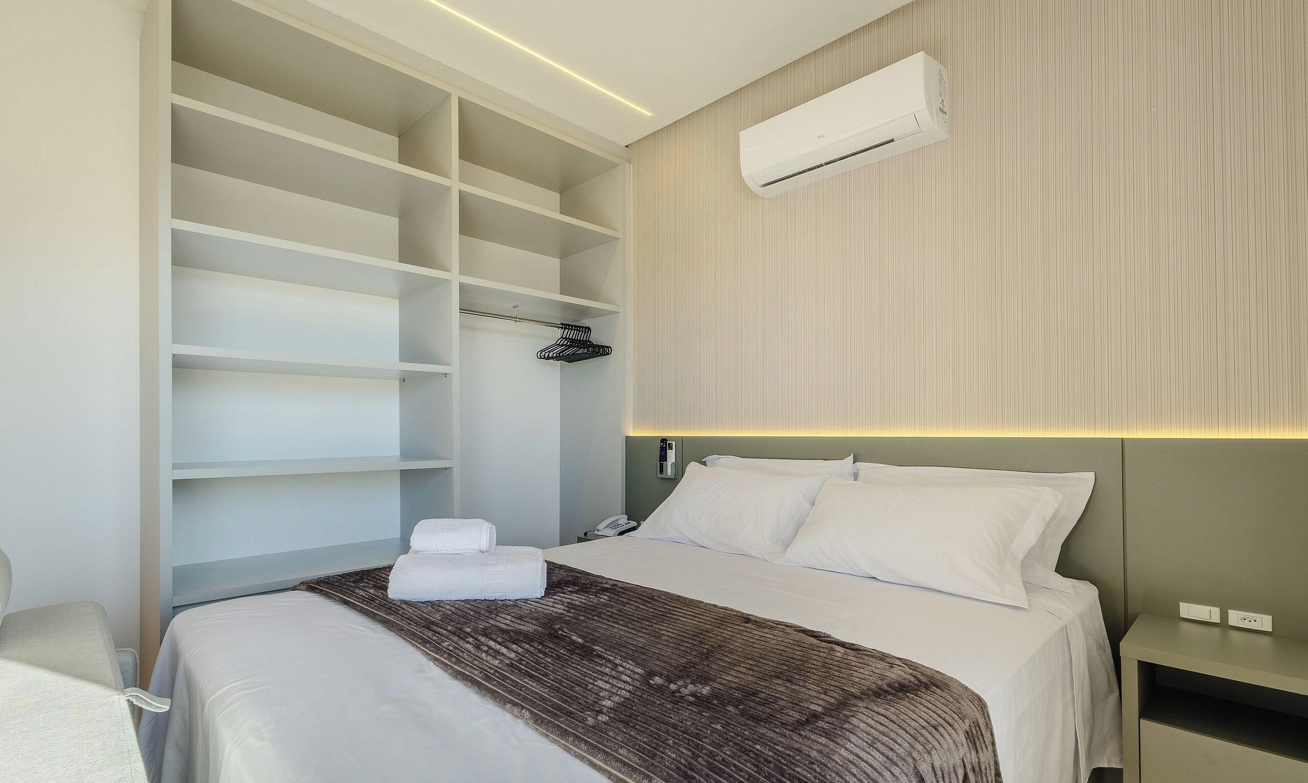 Habitación de hotel con cama, armario con estantes, mesita de noche y unidad de aire acondicionado en la pared.