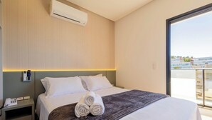 Ein Hotelzimmer mit einem Bett, einem Nachttisch, einer Wandklimaanlage und Blick auf Gebäude durch ein großes Fenster.