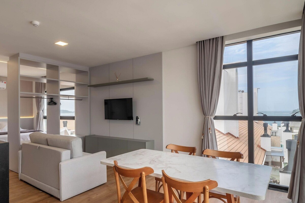 Una habitación de hotel moderna con una cama, un sofá, una mesa de comedor con sillas, un televisor montado en la pared y un ventanal con vistas a la ciudad.