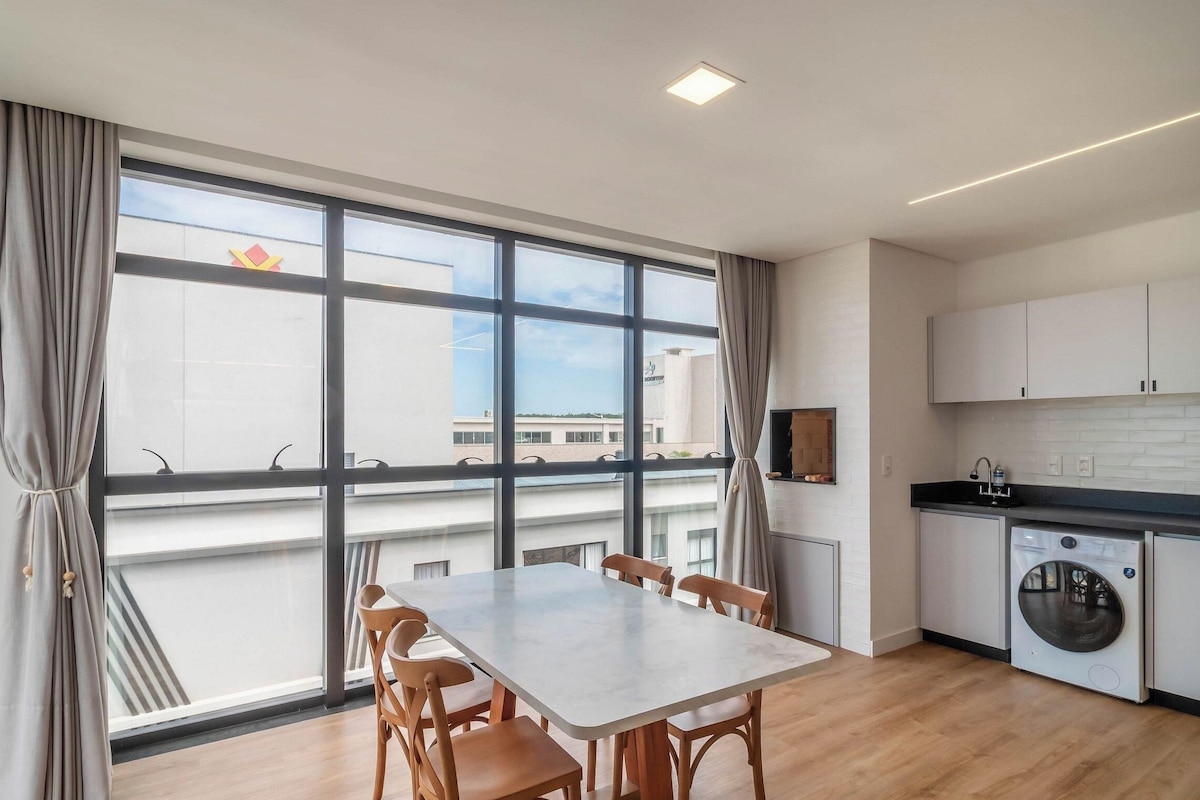 A modern kitchen with a built-in washing machine, a dining area with a table and chairs, and large windows offering a view of the cityscape.