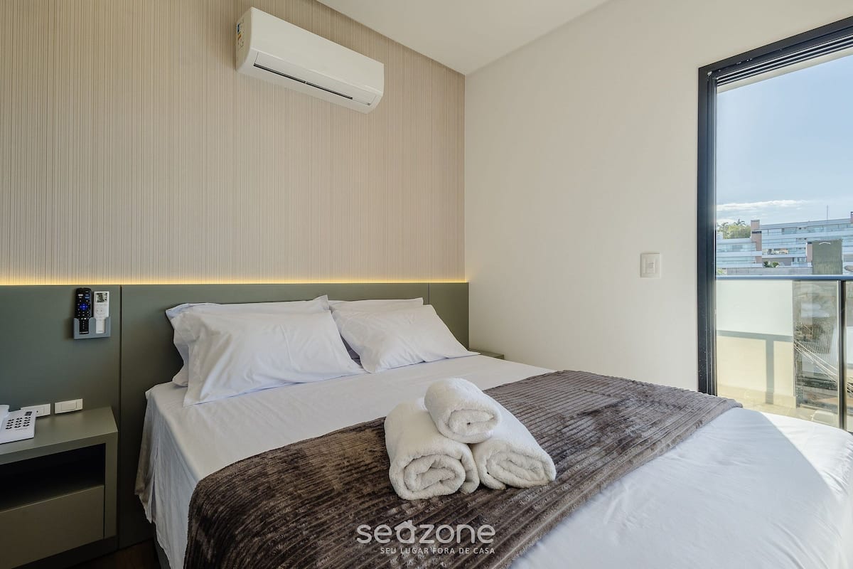 A hotel room with a bed, a bedside table with a phone, an air conditioner, and a window with a view.