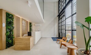 Un área de recepción con mostrador de madera, jardín vertical y zona de estar con sillas y mesas de madera.