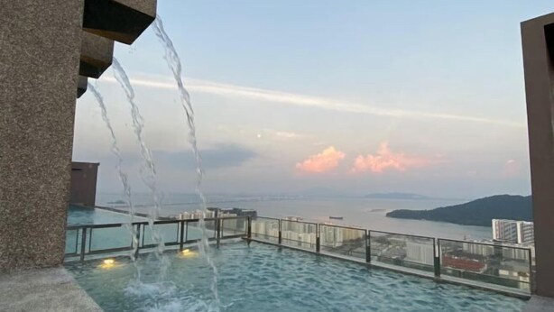 A rooftop infinity pool with water cascading over the edge, overlooking a cityscape and body of water. - Cozy 2BR Sky Pool & Panoramic Views 1-6 Guests (Gelugor)