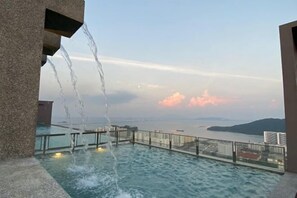 A rooftop infinity pool with water cascading over the edge, overlooking a cityscape and body of water. - Cozy 2BR Sky Pool & Panoramic Views 1-6 Guests (Gelugor)